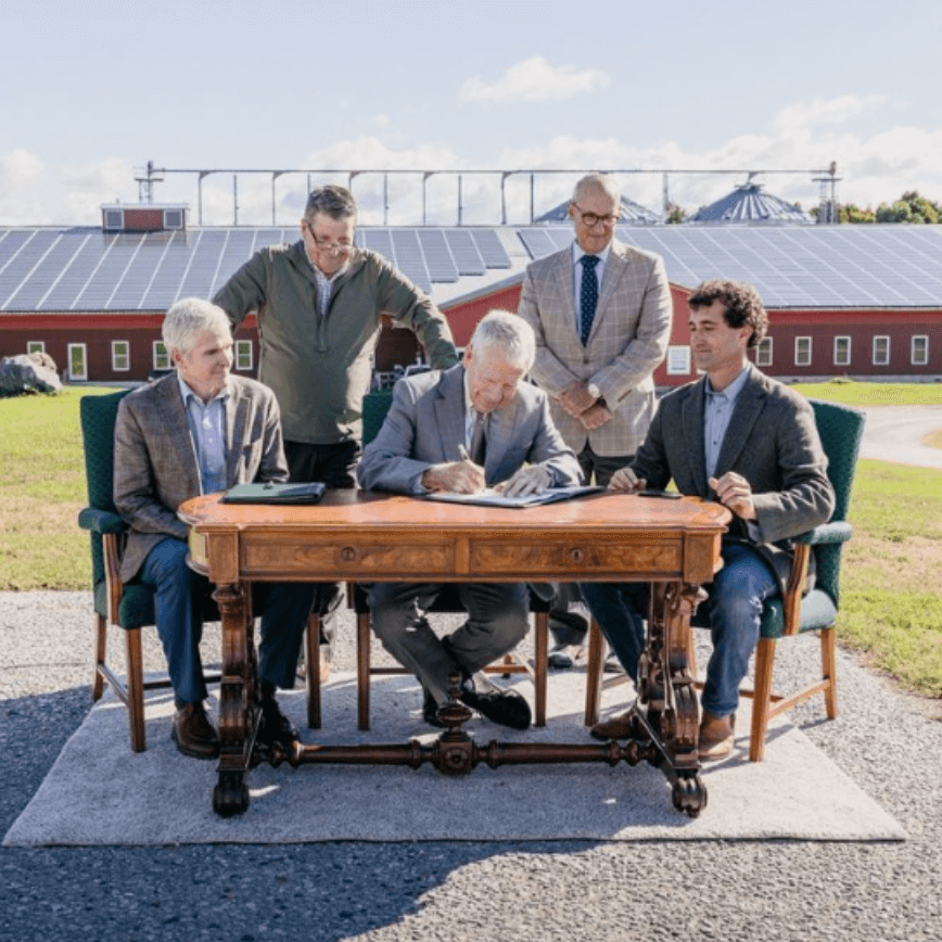 Five men gather at an antique writing desk in front of a barn to sign paperwork